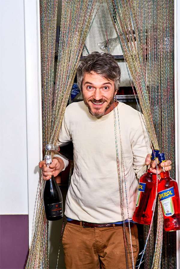 Giovanni Gentile grinning, carrying champagne and Aperol bottles out of kitchen